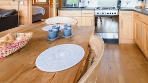 A cozy wooden cabin interior with a dining table set with blue cups and a woven placemat, a kitchenette with a stove and microwave, and a window overlooking a grassy landscape. - 2 bedroom accommodation in Linlithgow, near Edinburgh (Linlithgow, near Edinburgh)