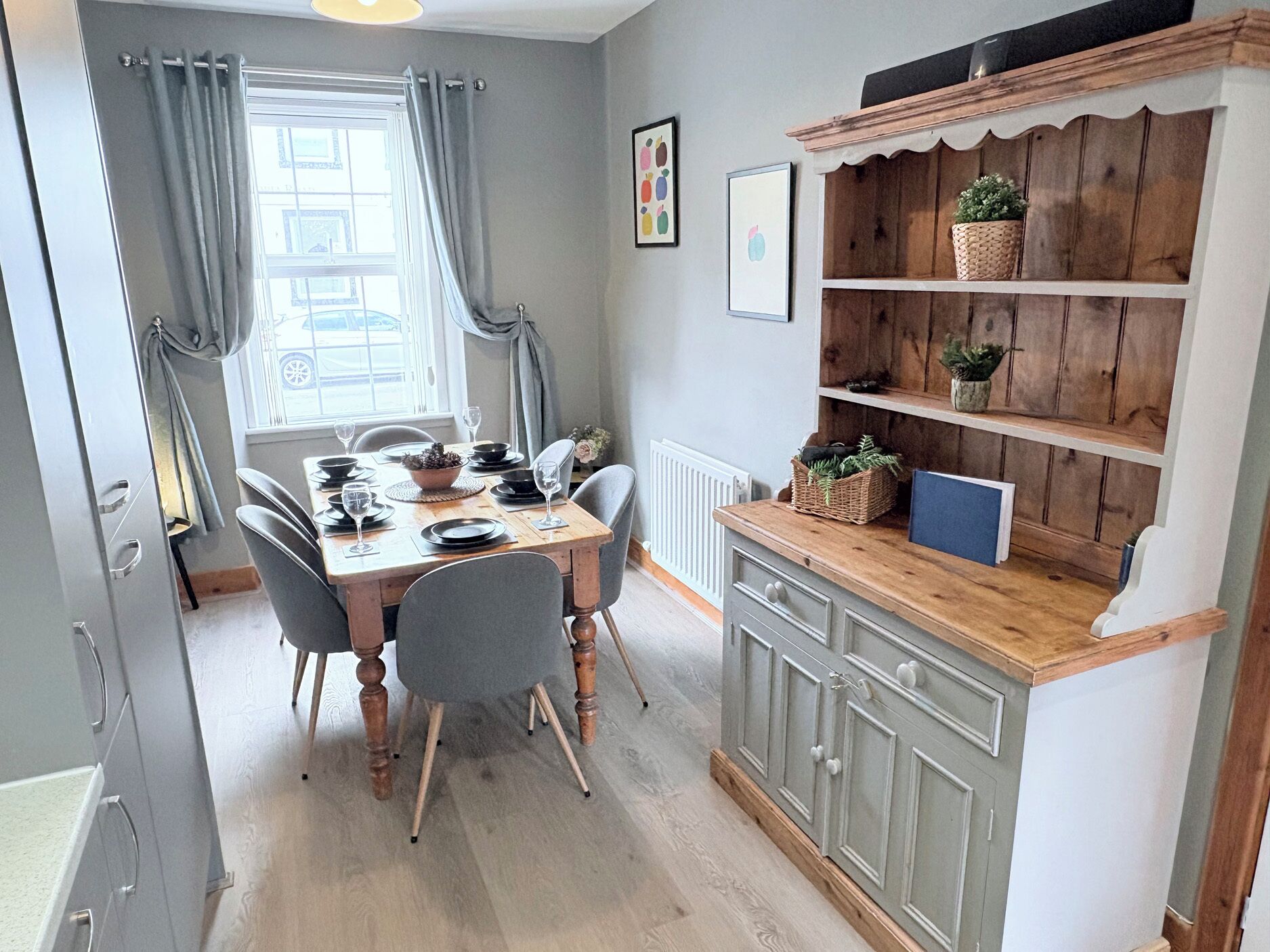 A dining area with a wooden table, grey chairs, and a kitchen with wooden shelves.