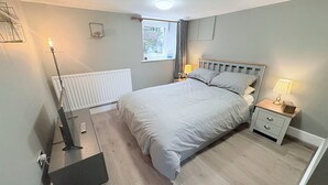 A bedroom with a bed, bedside tables, a radiator, a window, and a door. - The Oaks Cottage (Penrith)