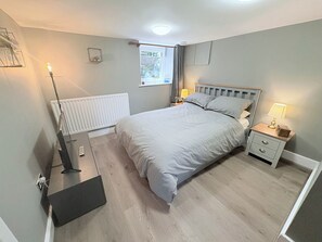A bedroom with a bed, bedside tables, a radiator, a window, and a door. - The Oaks Cottage (Penrith)