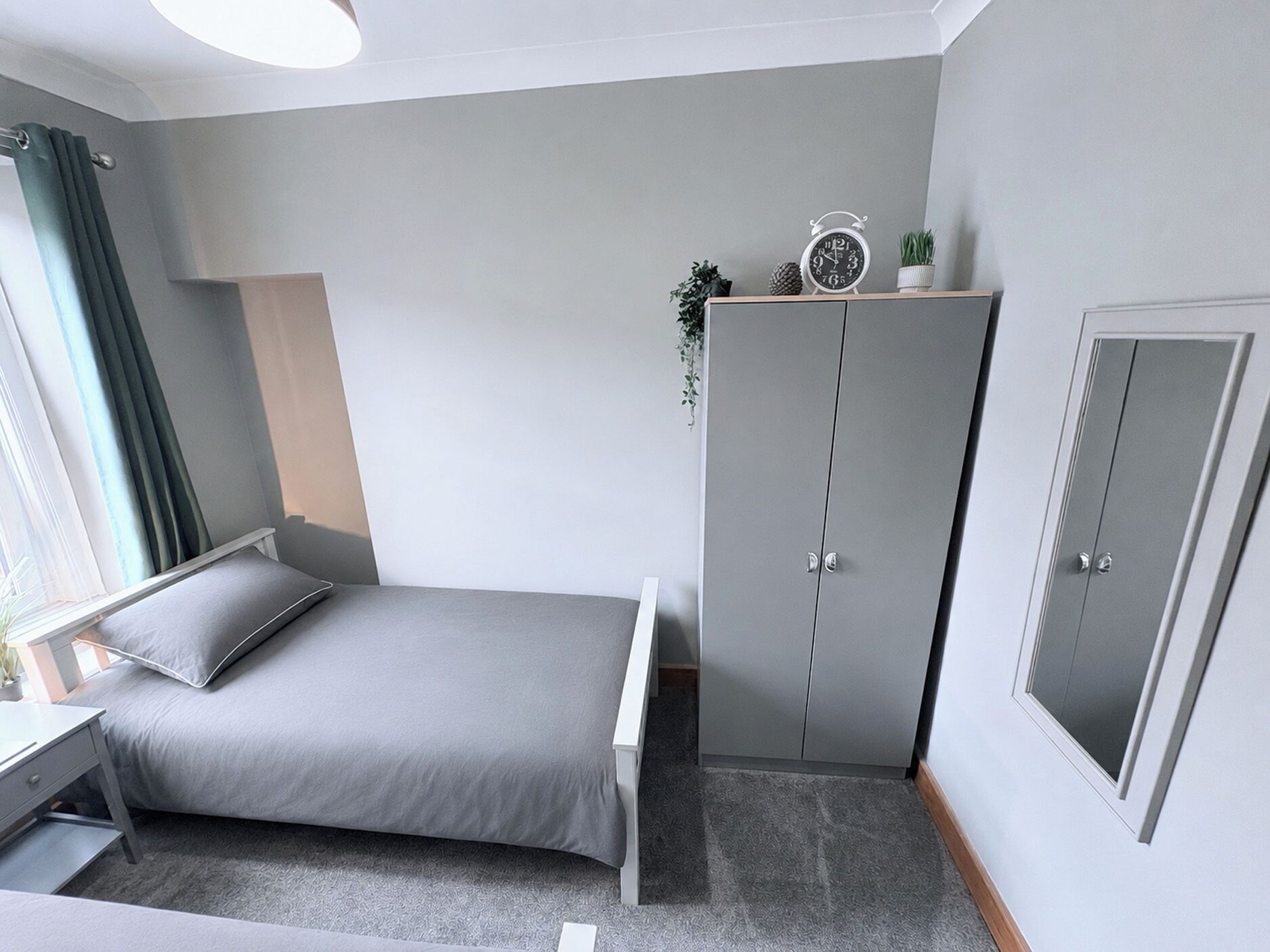 Ein Schlafzimmer mit Bett, Kleiderschrank, Nachttisch, einem Fenster mit Vorhängen und einer Wanduhr.