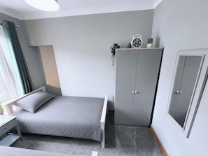 A bedroom with a bed, a wardrobe, a bedside table, a window with curtains, and a clock on the wall. - The Oaks Cottage (Penrith)