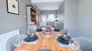A dining area with a wooden table set for four, flanked by grey chairs, and a kitchen in the background. - The Oaks Cottage (Penrith)