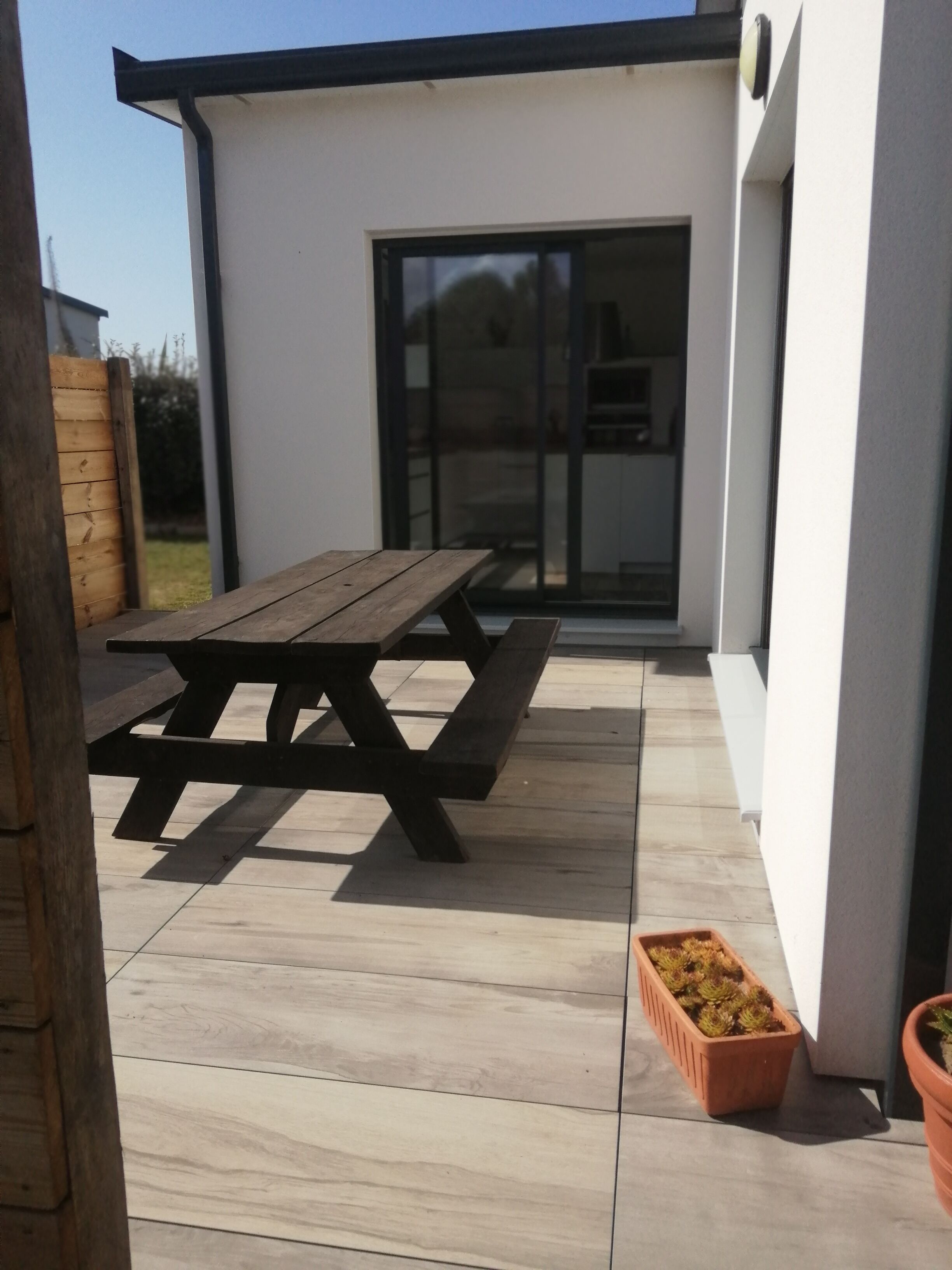 Een houten picknicktafel op een betegeld terras, met een wit gebouw op de achtergrond.