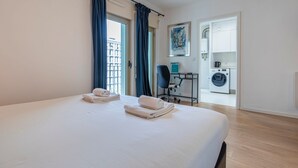 Habitación de hotel con una cama grande, un escritorio con silla, lavadora y una ventana con cortinas.
