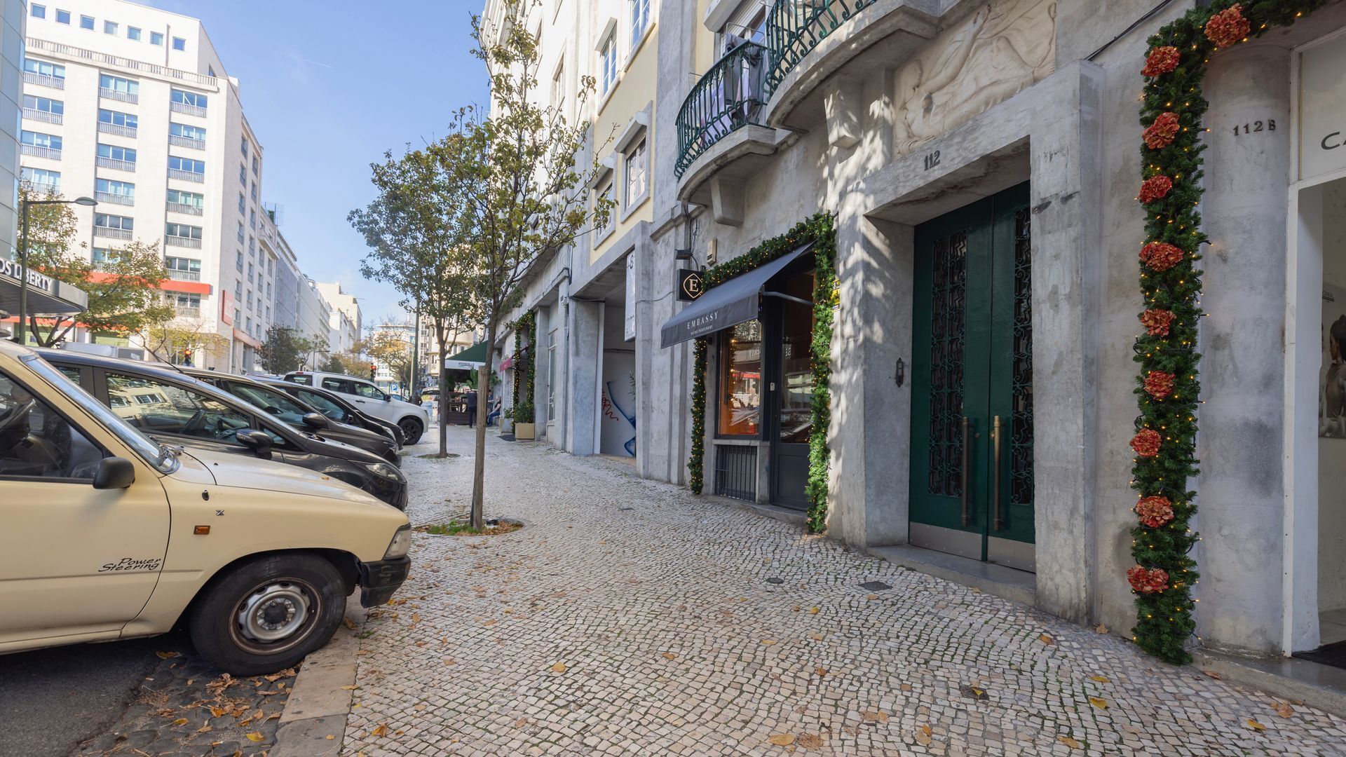 Eine Kopfsteinpflasterstraße mit geparkten Autos, ein Gebäude mit einer grünen Tür und einem Schild mit der Aufschrift 'EMBASSY'.