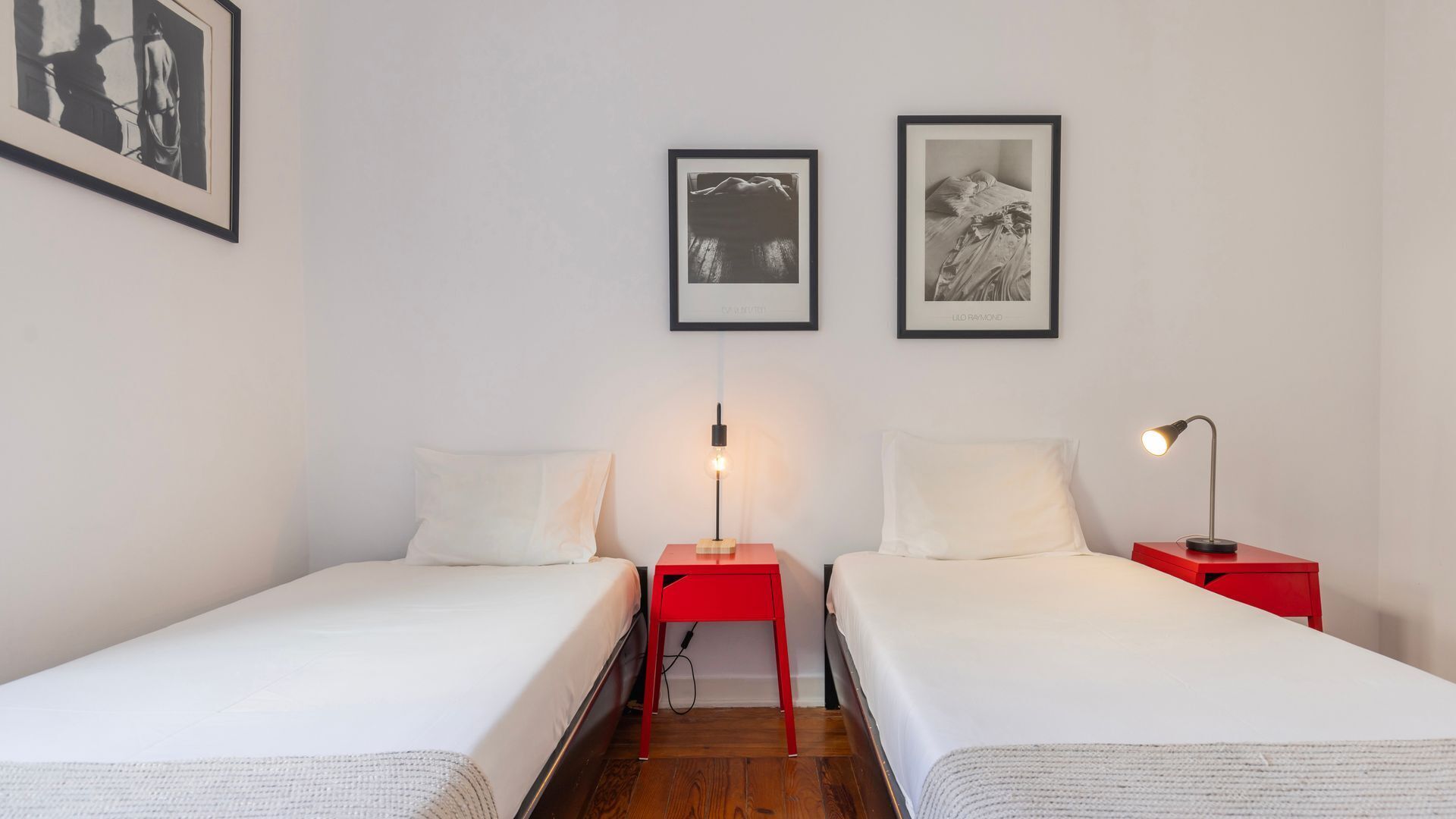 Een kamer met twee eenpersoonsbedden, elk met een rode nachtkastje en een tafellamp. Twee ingelijste foto’s hangen aan de muur.