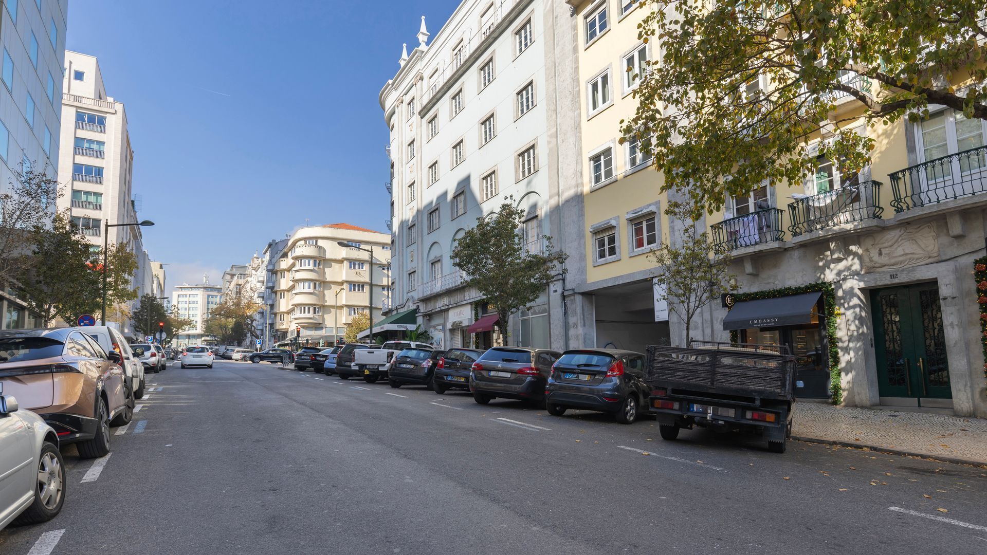 Una calle bordeada de coches aparcados y edificios, incluyendo un hotel con puerta verde y un cartel que dice 'EMBASSY'.