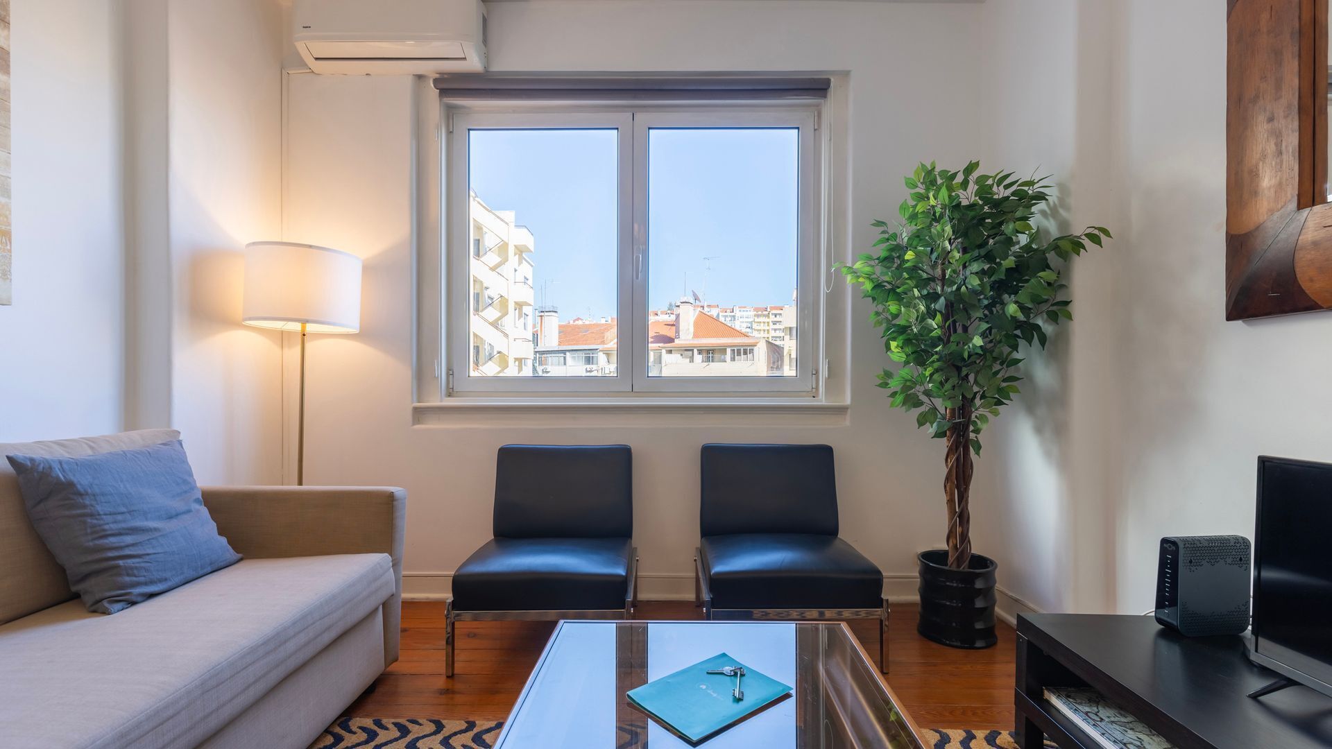Una sala de estar con sofá, dos sillones, una mesa de centro con un libro, una planta, televisor y ventana con vista a la ciudad.