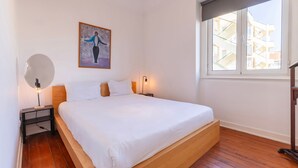 Un dormitorio minimalista con una cama grande, una mesita de noche con lámpara, un cuadro en la pared y una ventana con vistas a edificios.