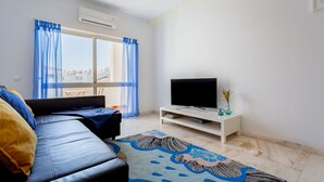 Un salon moderne avec un canapé, un téléviseur à écran plat posé sur un meuble blanc, un tapis à motifs et une vue par une fenêtre ornée de rideaux bleus.