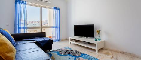 Ein modernes Wohnzimmer mit einer Couch, einem Flachbildfernseher auf einem weißen Schrank, einem gemusterten Teppich und Blick durch ein Fenster mit blauen Vorhängen.