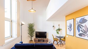 A modern living room with a large TV, a sofa, a dining area with chairs, and a potted plant.