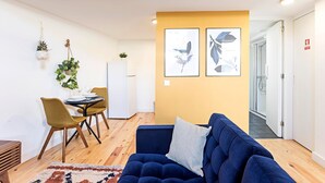 A modern living room with a blue sofa, a dining area with a table and chairs, and a kitchen with a white refrigerator.