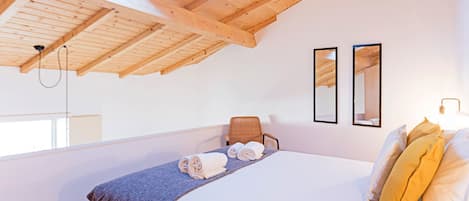 A bedroom with a high wooden ceiling, a bed with rolled towels, a chair, and two framed mirrors on the wall.