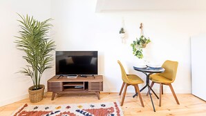 A cozy living area with a flat-screen TV on a wooden stand, a potted plant, a small dining table with two chairs, and a hanging plant decoration.