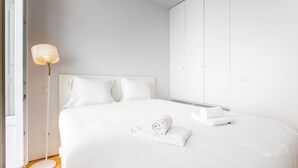 Una cama bien tendida con sábanas blancas y dos almohadas, una lámpara de noche y un conjunto de armarios blancos.