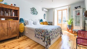 A bedroom with a wooden floor, a bed with a floral bedspread, a wooden wardrobe, a blue lamp, and a potted plant. - Lisbon Heart Apartments - Yellow, by LovelyStay (Lisboa)