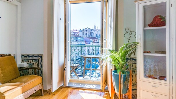 A room with a wooden floor, a sofa, a potted plant, a cabinet, and a view of a cityscape through an open door. - Lisbon Heart Apartments - Yellow, by LovelyStay (Lisboa)