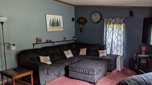 A living room with a sectional sofa, a small wooden table, a floor lamp, a clock, and a television. - Hackert Lake House near Ludington! (Scottville)