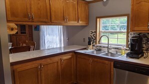 A kitchen with wooden cabinets, a stainless steel dishwasher, a coffee maker, and a window with a view of greenery. - Hackert Lake House near Ludington! (Scottville)