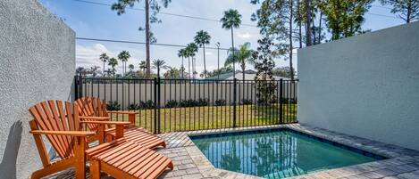 A pool area with a wooden deck and chairs, surrounded by a fence and palm trees.