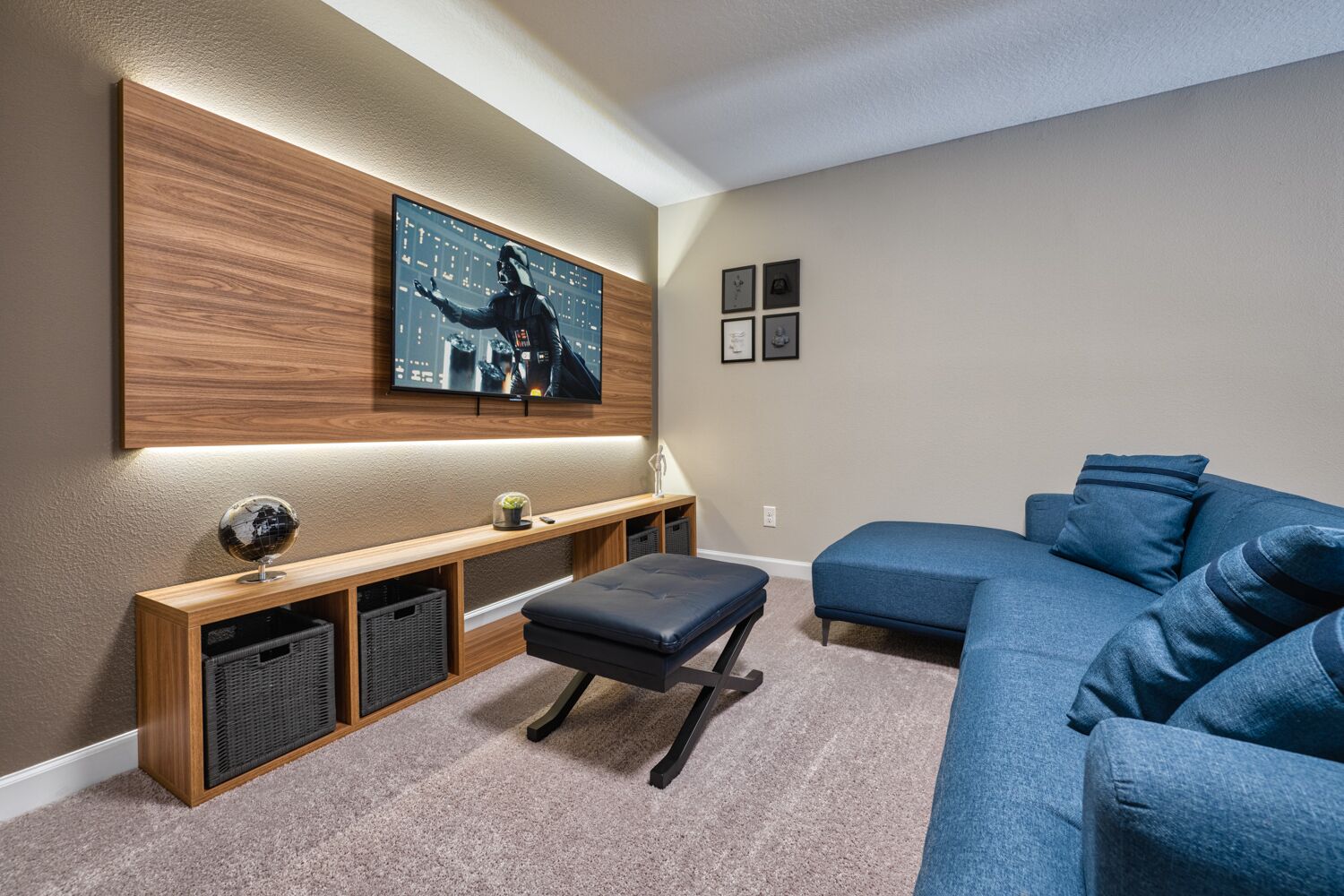 A modern living room with a large flat-screen TV mounted on a wooden panel, a blue sectional sofa, a small ottoman, and a wooden console table with storage baskets.