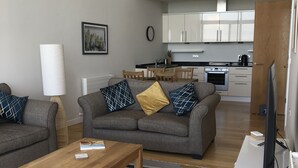 A modern living room with a sofa, coffee table, and a TV. A kitchen is visible in the background. - Penthouse apartment with stunning sea view Westward Ho! (Westward Ho!)