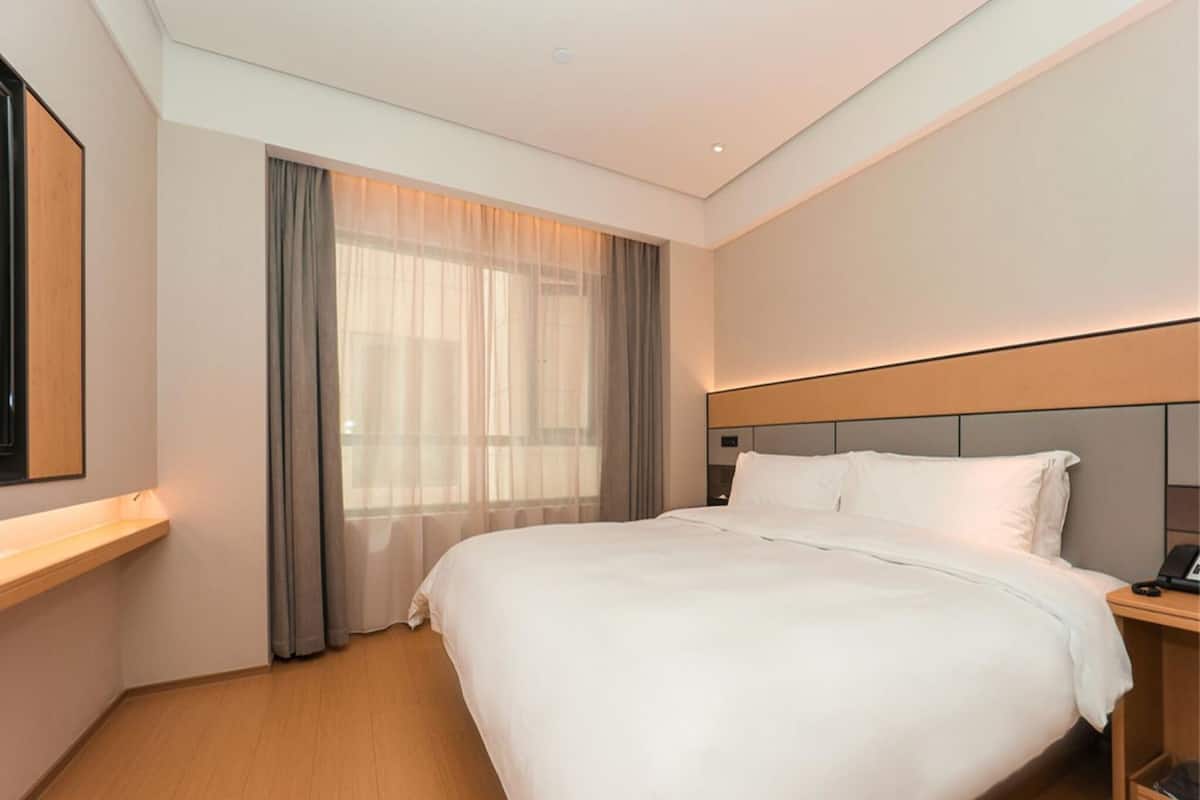A hotel room with a large bed, a bedside table with a phone, a window with curtains, and a wall-mounted TV.