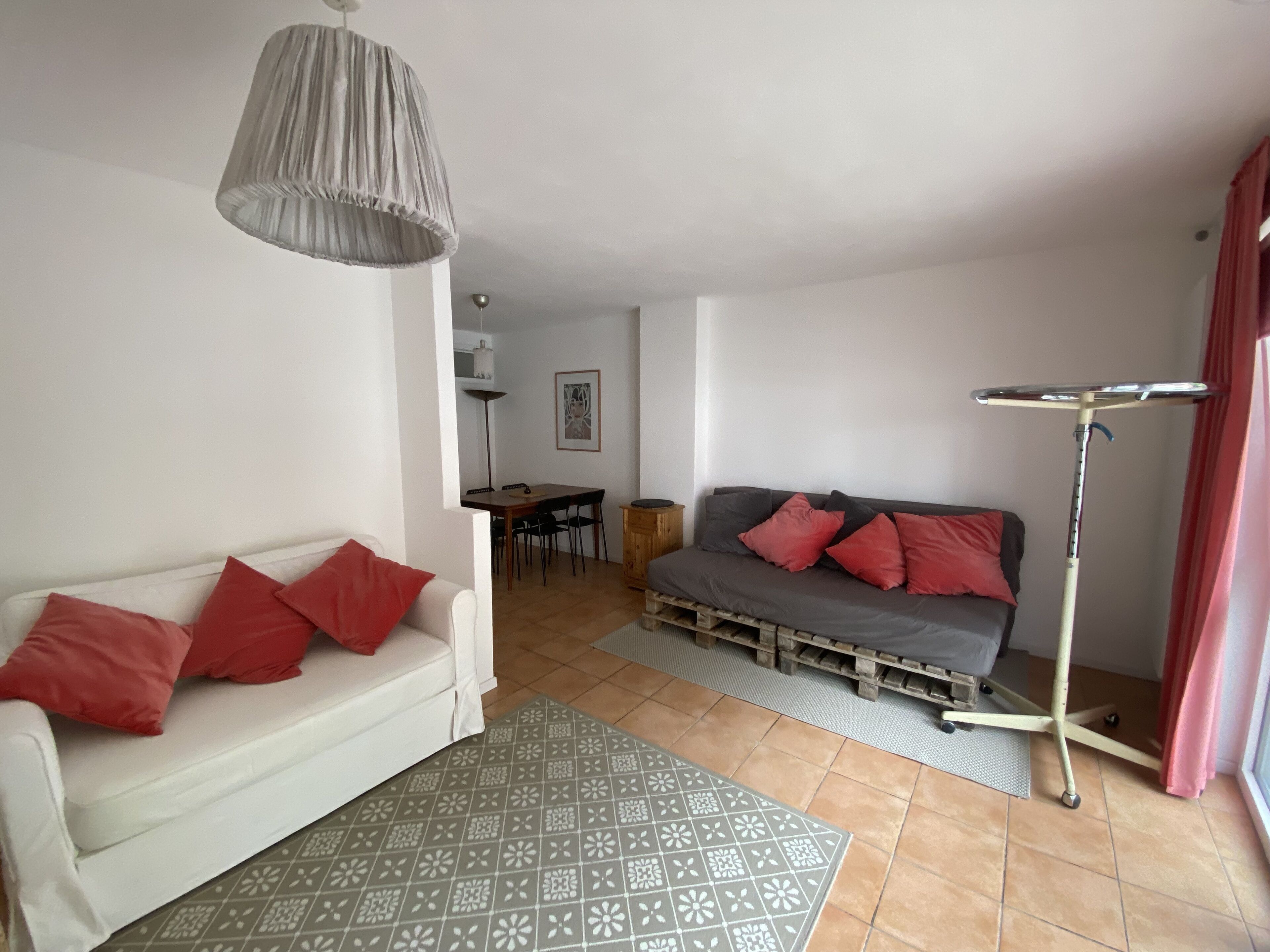 A living room with a white sofa, red cushions, a grey sofa with red cushions, a dining table with chairs, and a floor lamp.
