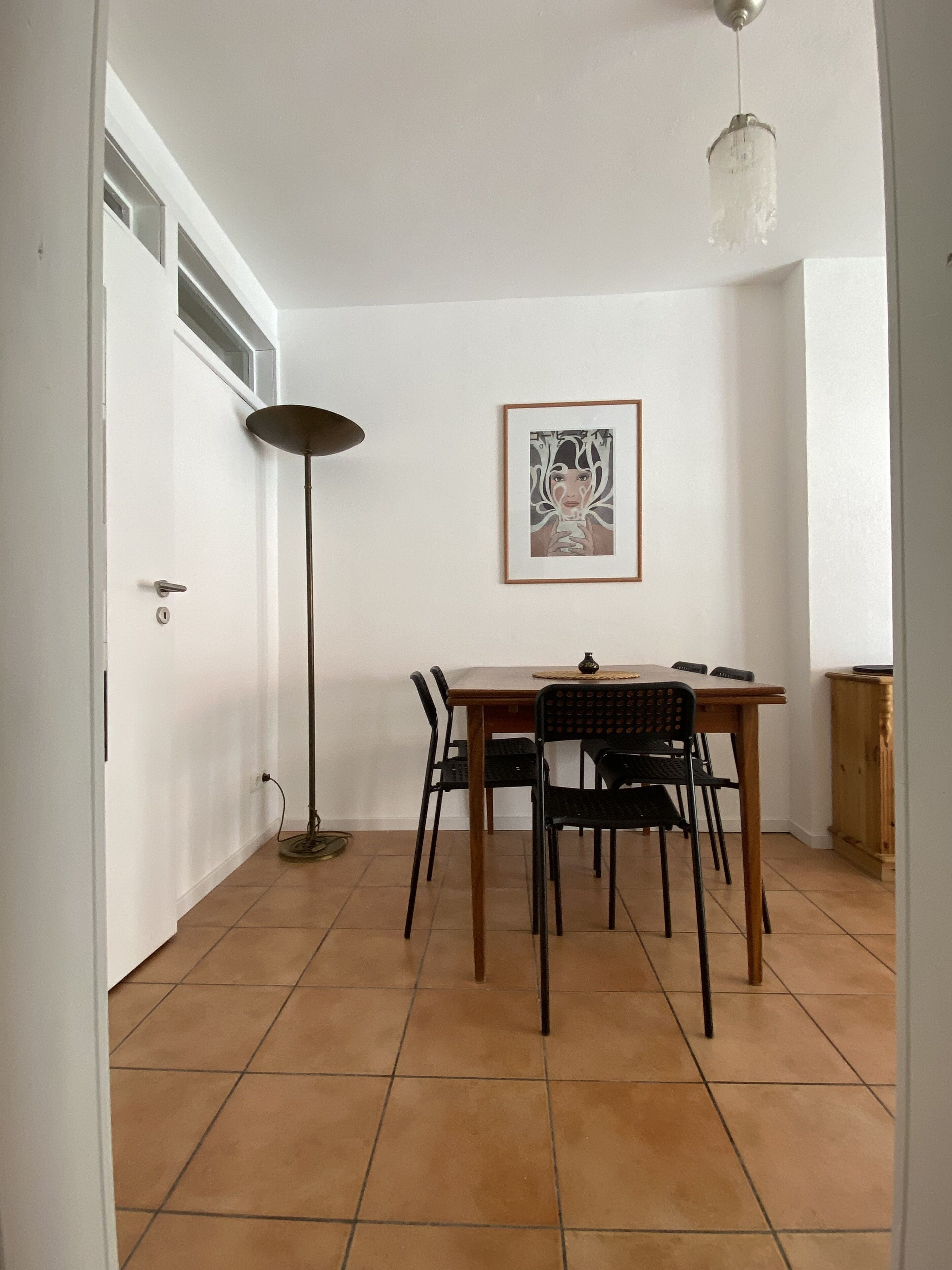 Een eethoek met een houten tafel en stoelen, een vloerlamp en een kunstwerk in een lijst aan de muur.
