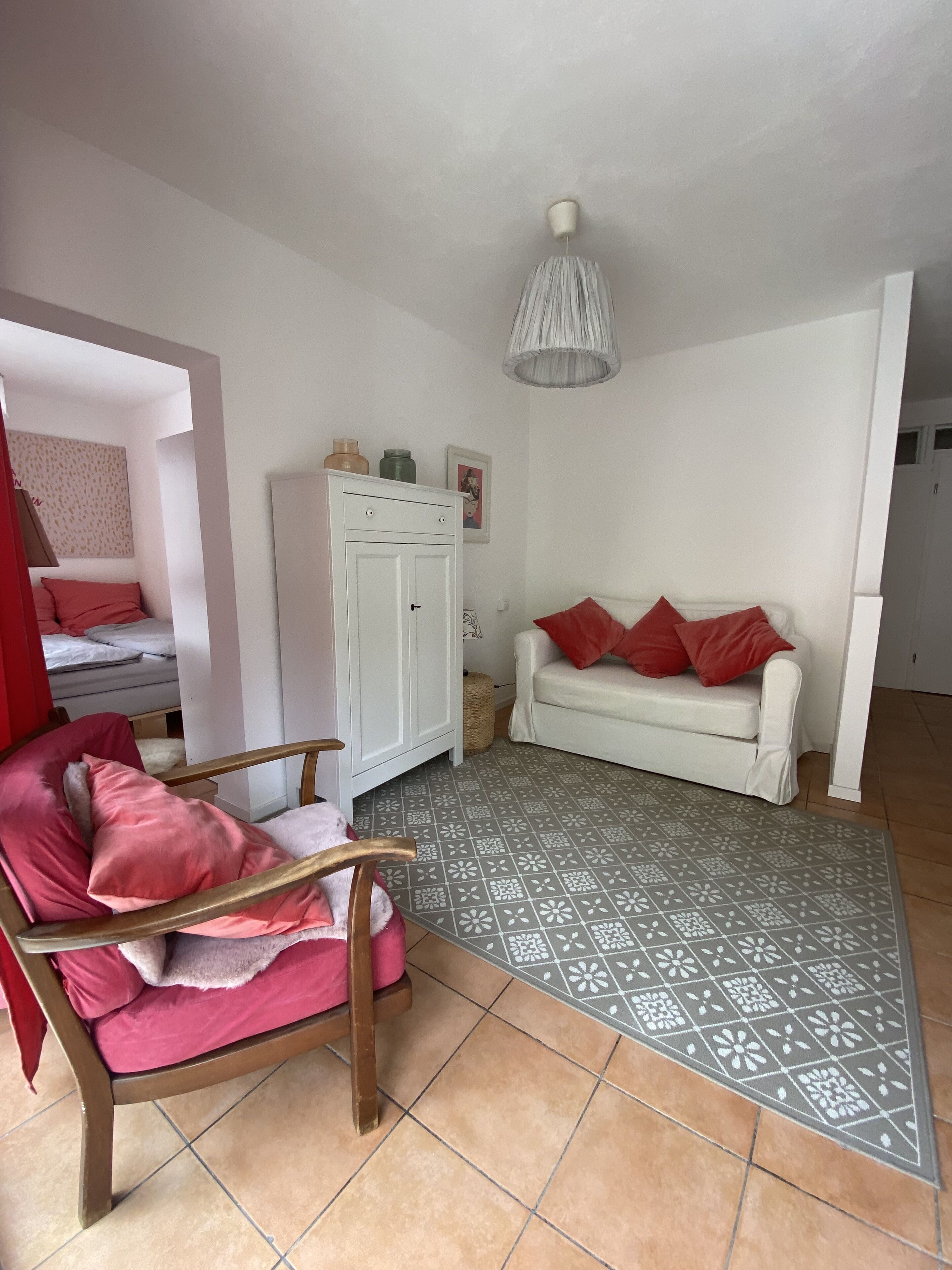 A room with a wooden chair, a white sofa with red cushions, a white wardrobe, and a patterned rug.