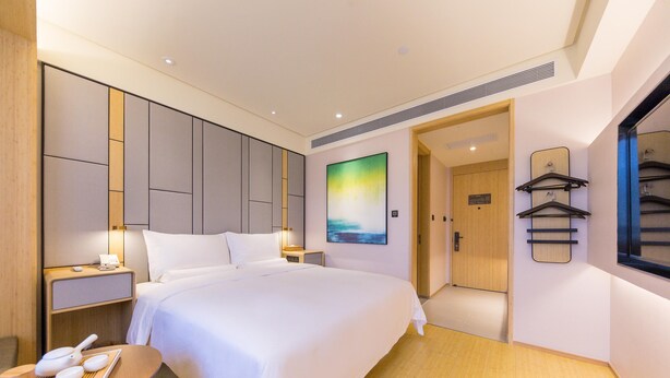 Habitación de hotel moderna con una cama grande, mesitas de noche, una pequeña mesa con bandeja, un cuadro en la pared y un espejo con perchero.