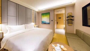 Una habitación de hotel con una cama grande, un sofá, una mesita con una tetera, un cuadro y una puerta que da a otra habitación.