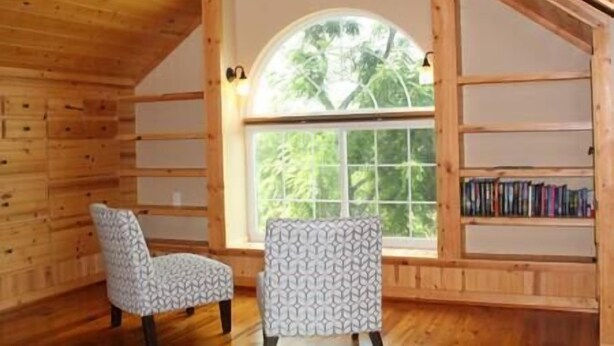 Una habitación de cabaña acogedora, con interior de madera, un ventanal con vistas a árboles, una silla estampada y una estantería.