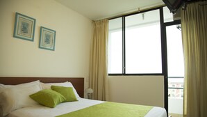 A hotel room with a bed, two framed pictures on the wall, a window with curtains, and a view of buildings outside. - Hotel RC Tonsupa (Tonsupa)