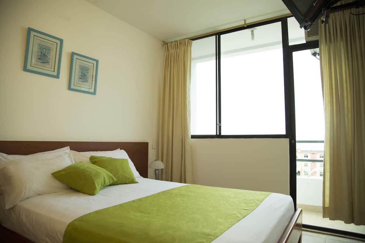 Quarto de hotel com uma cama, duas fotografias emolduradas na parede, uma janela com cortinas e vista para edifícios.