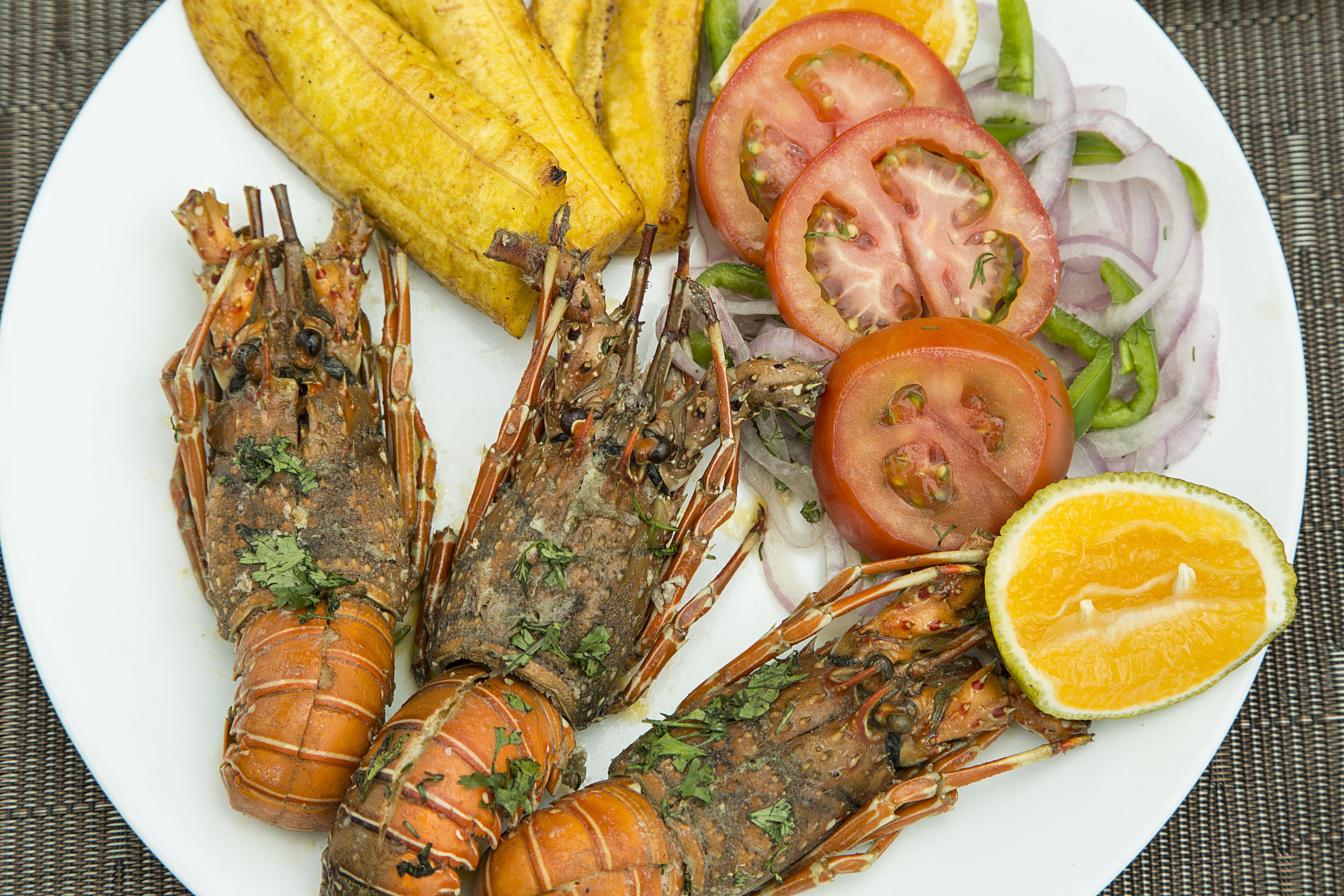 A plate with grilled lobster, fried plantains, sliced tomatoes, onions, and a slice of lime.