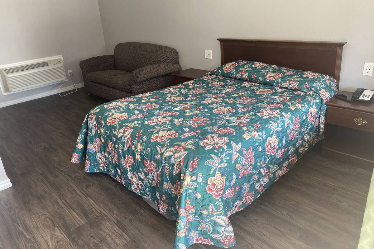 A bedroom with a floral bedspread, a wooden nightstand, a chair, and a wall-mounted heater.