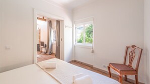Quarto com cama, cadeira, janela e porta que dá para outro cômodo.