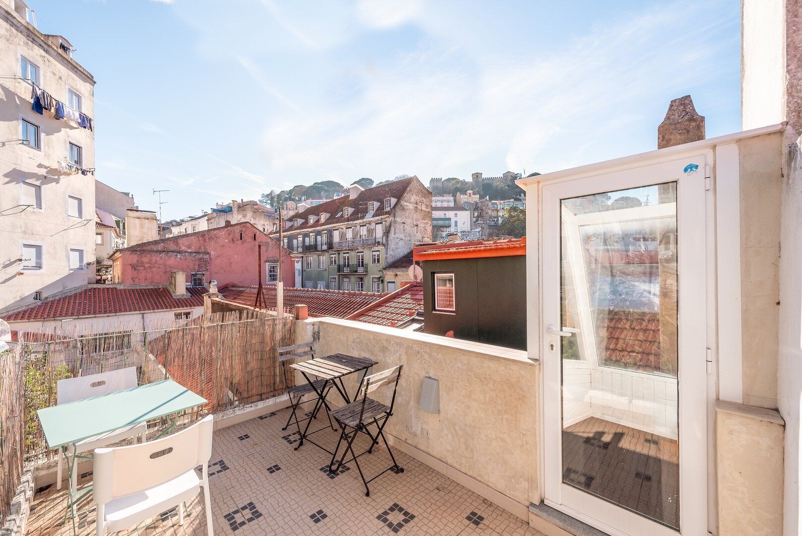 Un balcón con mesa y sillas, vista a un paisaje urbano con edificios históricos, y cielo despejado.