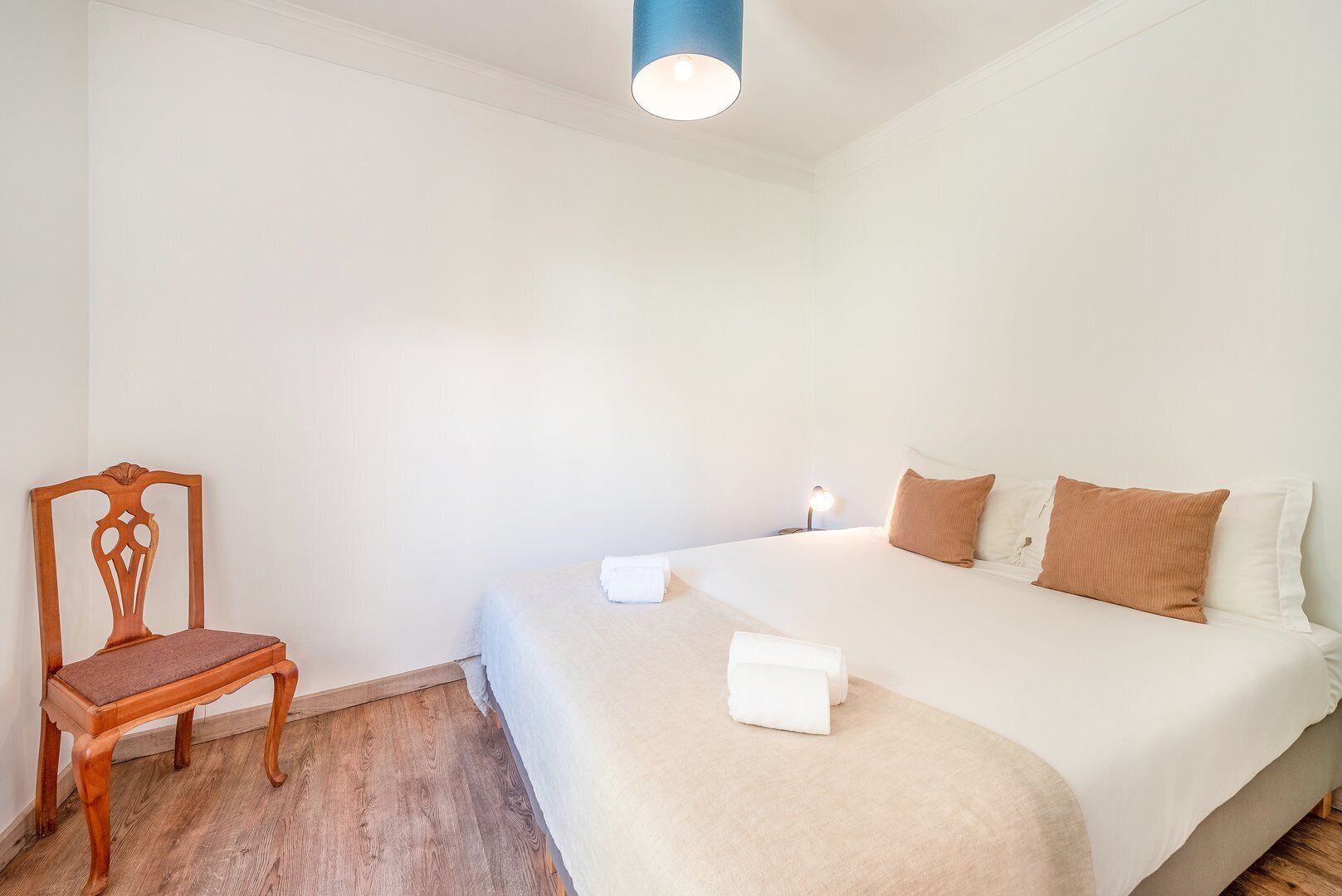 Un dormitorio con una silla de madera, una cama con almohadas blancas y marrones, y una lámpara de noche.
