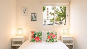 A bedroom with a bed, two bedside lamps, a window with a view of buildings, and two framed botanical prints on the wall. - GuestReady - Great 3BDR apartment in Oeiras! (Oeiras)