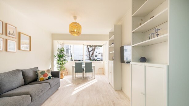 A modern living room with a grey sofa, a dining table with chairs, a large window, and a white cabinet with shelves. - GuestReady - Great 3BDR apartment in Oeiras! (Oeiras)
