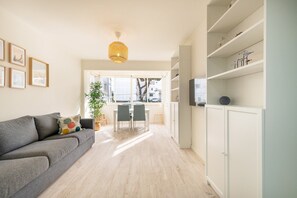 A modern living room with a grey sofa, a dining table with chairs, a large window, and a white cabinet with shelves. - GuestReady - Great 3BDR apartment in Oeiras! (Oeiras)