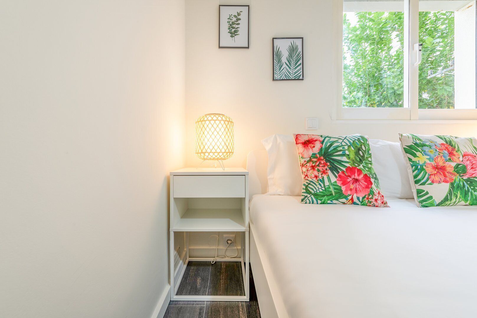 Un dormitorio con una cama, una mesita de noche, una lámpara y dos impresiones botánicas enmarcadas en la pared.