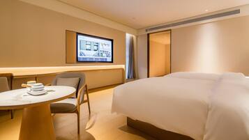 A hotel room with a bed, a desk with a chair, a television mounted on the wall, and a window with curtains.