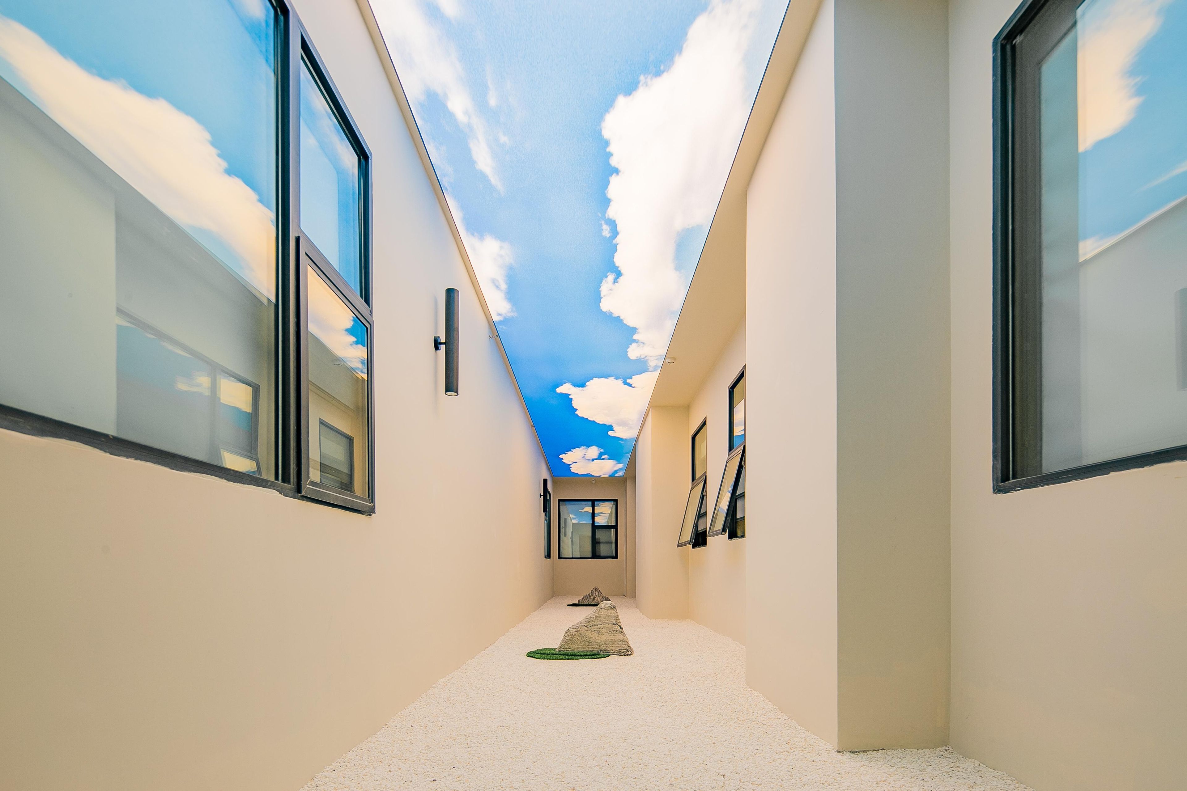 Un couloir étroit dont le plafond est peint pour ressembler à un ciel nuageux, avec de grandes fenêtres de chaque côté et un petit tapis sur le sol.