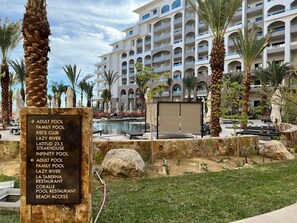 Un complexe hôtelier doté d’une piscine, de palmiers et d’un panneau indiquant les équipements disponibles, tels que des piscines pour adultes et pour familles, un restaurant servant des steaks et un restaurant situé au bord de la piscine.