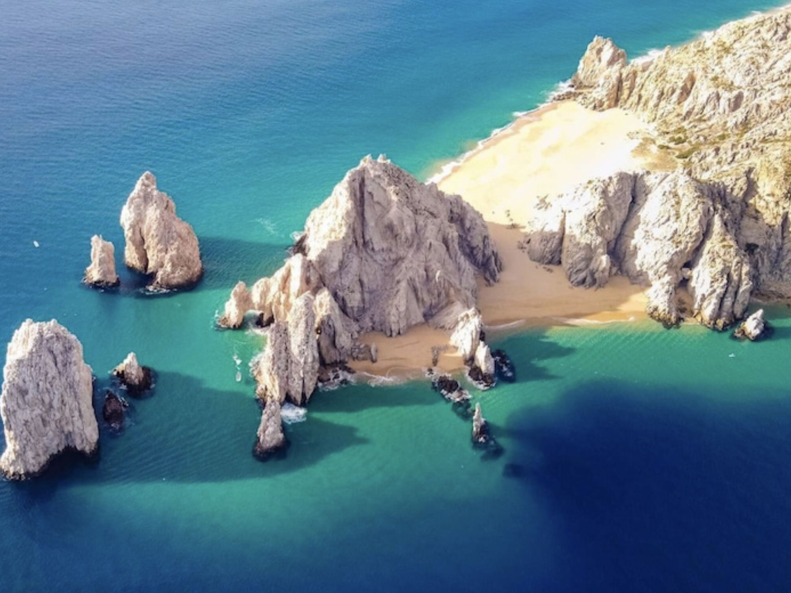 An aerial view of a coastal area with rocky formations, clear blue waters, and a sandy beach.